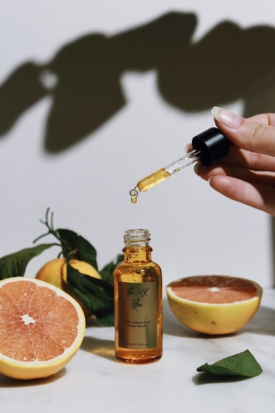A high-end product hero shot featuring a 10% Vitamin C + E face serum bottle with fresh grapefruit segments and a dropper in motion.